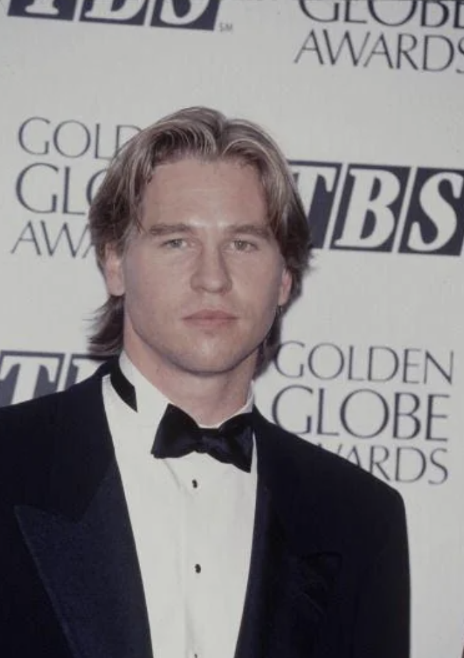 A man wearing a black tuxedo with a black bow tie stands in front of a backdrop with the printed text "Golden Globe Awards" and "TBS." He has medium-length, light brown hair and is looking straight at the camera.