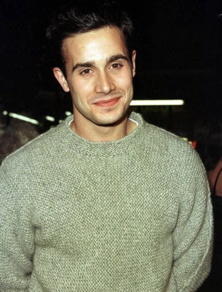 A man with short, dark hair, wearing a light gray knit sweater, smiles warmly at the camera. He is in an indoor setting with blurred background elements and appears to be in casual attire.