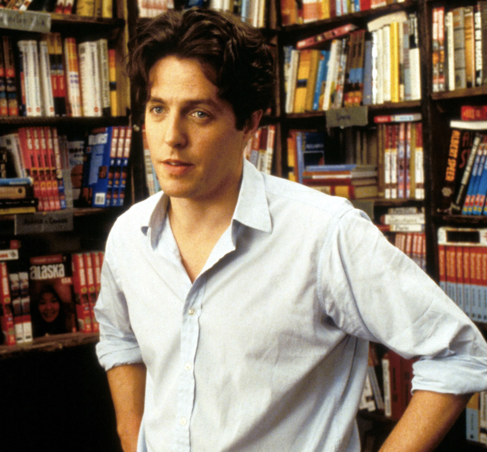 A person with dark, wavy hair is standing in a bookstore with shelves full of books and magazines in the background. They are wearing a light blue shirt with the sleeves rolled up and looking slightly to the side with a thoughtful expression.