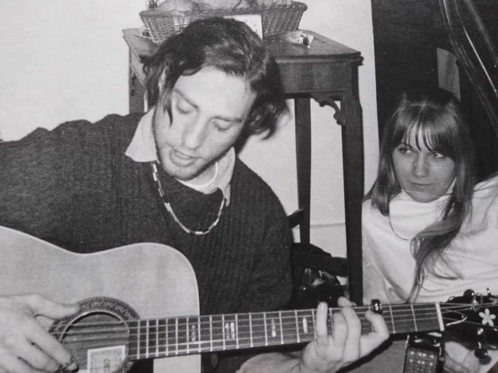 Black and white photo of a man and a woman. The man, with mid-length hair, is sitting and playing an acoustic guitar. The woman, with long hair and bangs, is seated behind him, looking at him attentively. They are indoors, and a vintage table is visible in the background.