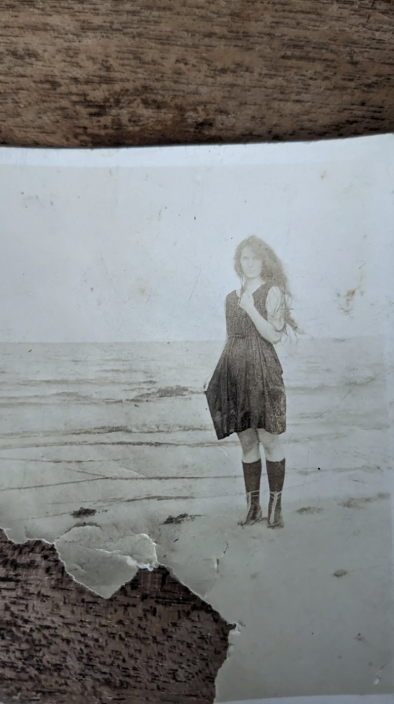 A sepia-toned photograph of a young girl standing on a beach. She is wearing a dark dress and knee-high socks, with her long hair flowing in the wind. The ocean waves can be seen in the background, and the photo shows some signs of aging and wear.