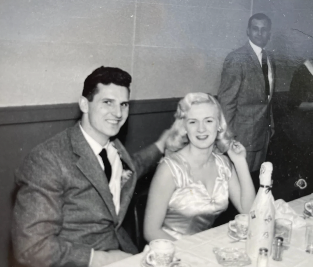 Black and white photo of a man and woman sitting at a table, both smiling. The man is wearing a suit and tie, and the woman is in a satin dress. The table has teacups, saucers, and a bottle. Another man in a suit stands in the background.
