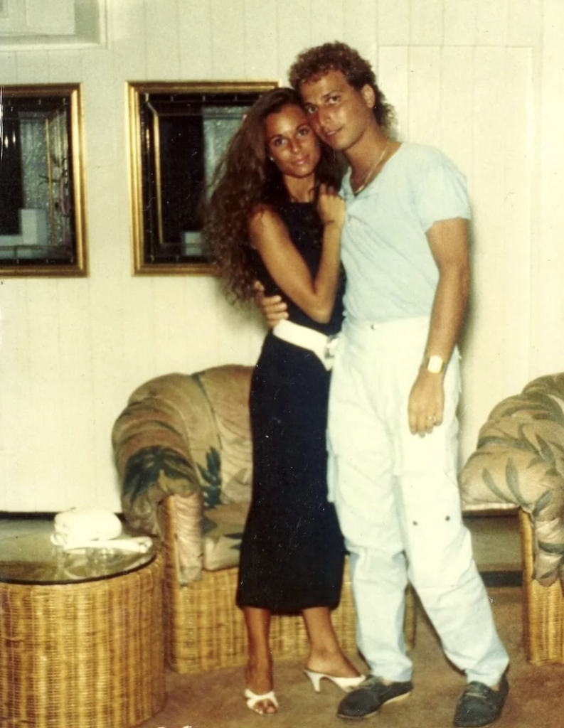 A couple stands closely together in a living room. The woman, with long wavy hair and a black dress, is embraced by the man, who is wearing a light blue shirt and white pants. They are standing next to a wicker chair and table, with framed pictures on the wall behind them.