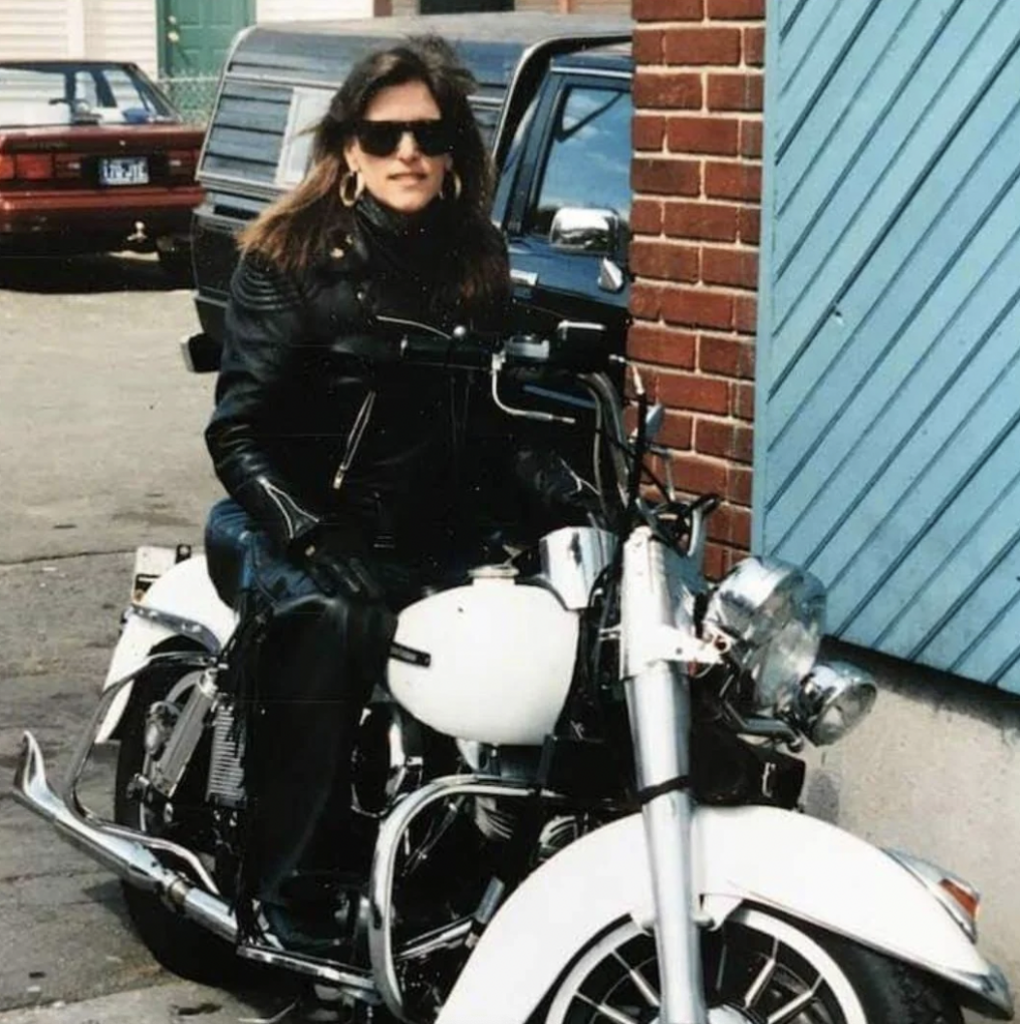 A person dressed in black leather jacket and gloves, wearing sunglasses, is sitting on a white motorcycle parked next to a light blue, wooden wall in an urban area. A black vehicle and a brick wall are visible in the background.
