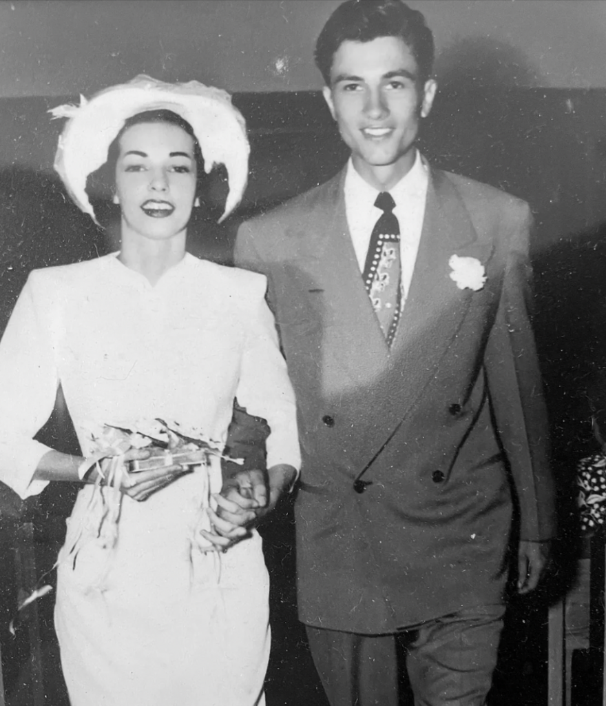 A black-and-white photo of a smiling couple dressed in formal attire, seemingly from a mid-20th century event. The woman is wearing a white dress and a stylish hat, holding a small bouquet. The man is in a light-colored suit with a patterned tie and boutonnière.