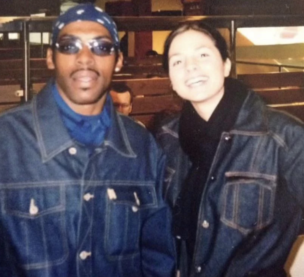 Two people standing side by side in a casual setting. The person on the left is wearing sunglasses, a bandana, and a denim jacket, while the person on the right is smiling, wearing a black scarf and denim jacket. They are indoors with a blurred background of seats.