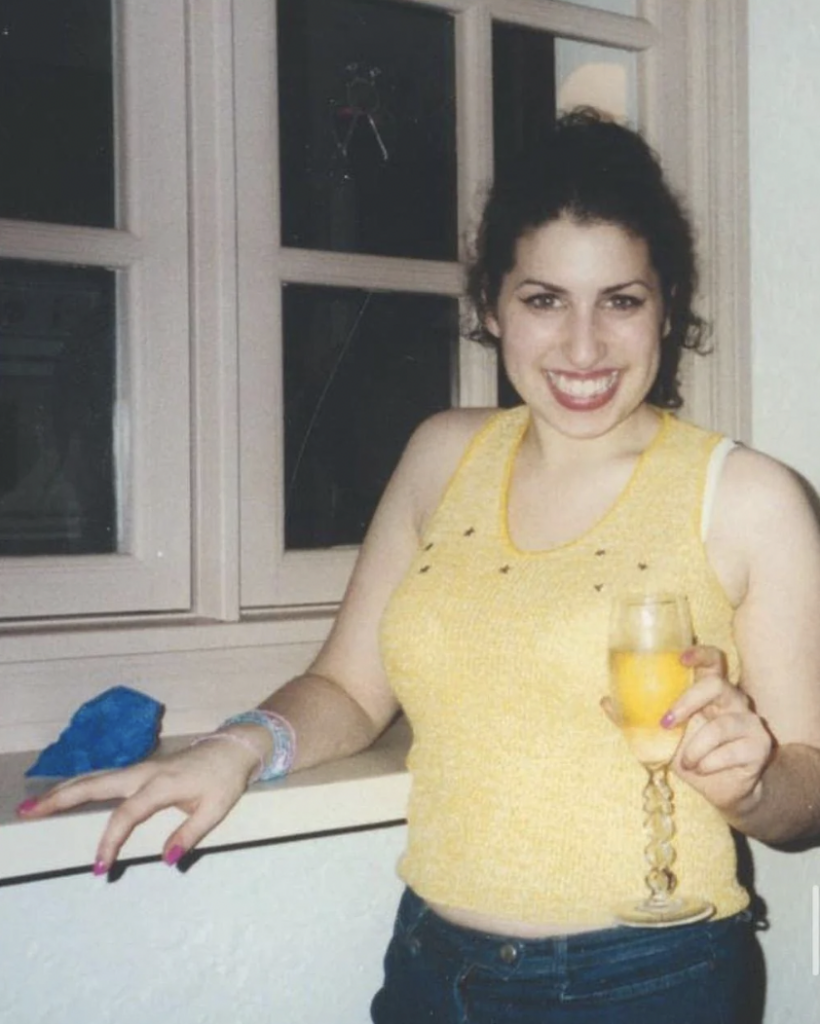 A person with dark hair stands indoors smiling at the camera. They are wearing a yellow sleeveless top and blue jeans and are holding a filled champagne flute in one hand. They are standing by a window with light-colored frames.