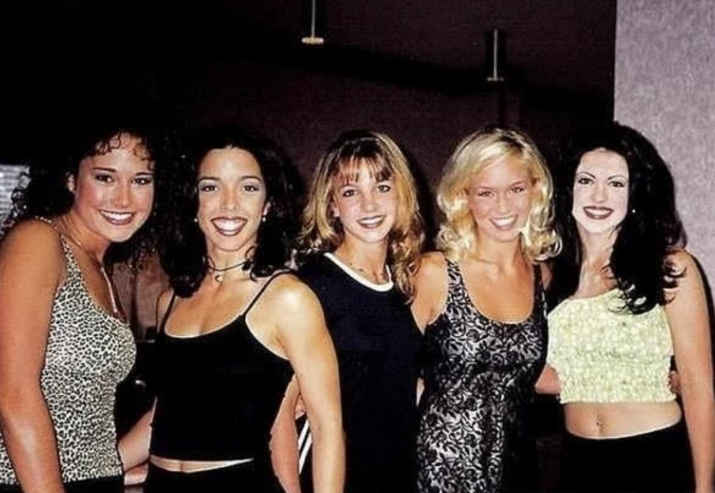 A group of five women are smiling and posing together for a photo. They are dressed in various outfits, with most wearing sleeveless tops. The background appears to be an indoor setting with dim lighting.