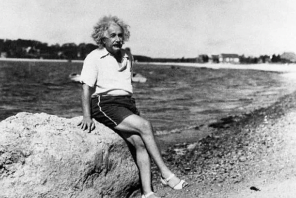 A black-and-white photograph of a man with wild, white hair and a mustache, wearing a white shirt and shorts, sitting casually on a large rock near a body of water. The shoreline and distant buildings are visible in the background. He appears relaxed and thoughtful.