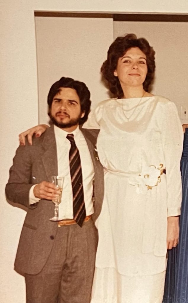 A woman in a white dress with long sleeves stands beside a man in a gray suit and black striped tie. The man holds a champagne glass, and both appear smiling. The woman has her right arm around the man’s shoulder, and they are indoors with white walls in the background.