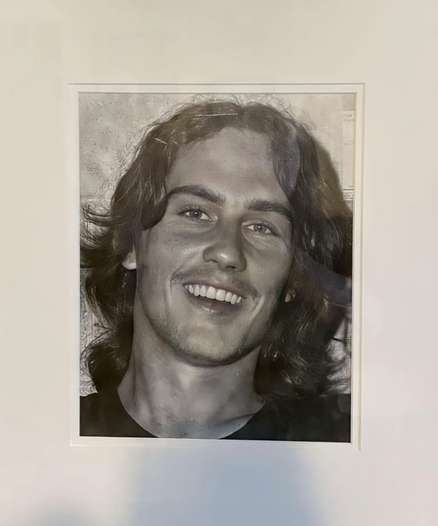 A black-and-white portrait of a young man with shoulder-length wavy hair smiling warmly. He is framed against a plain background, wearing a dark shirt.