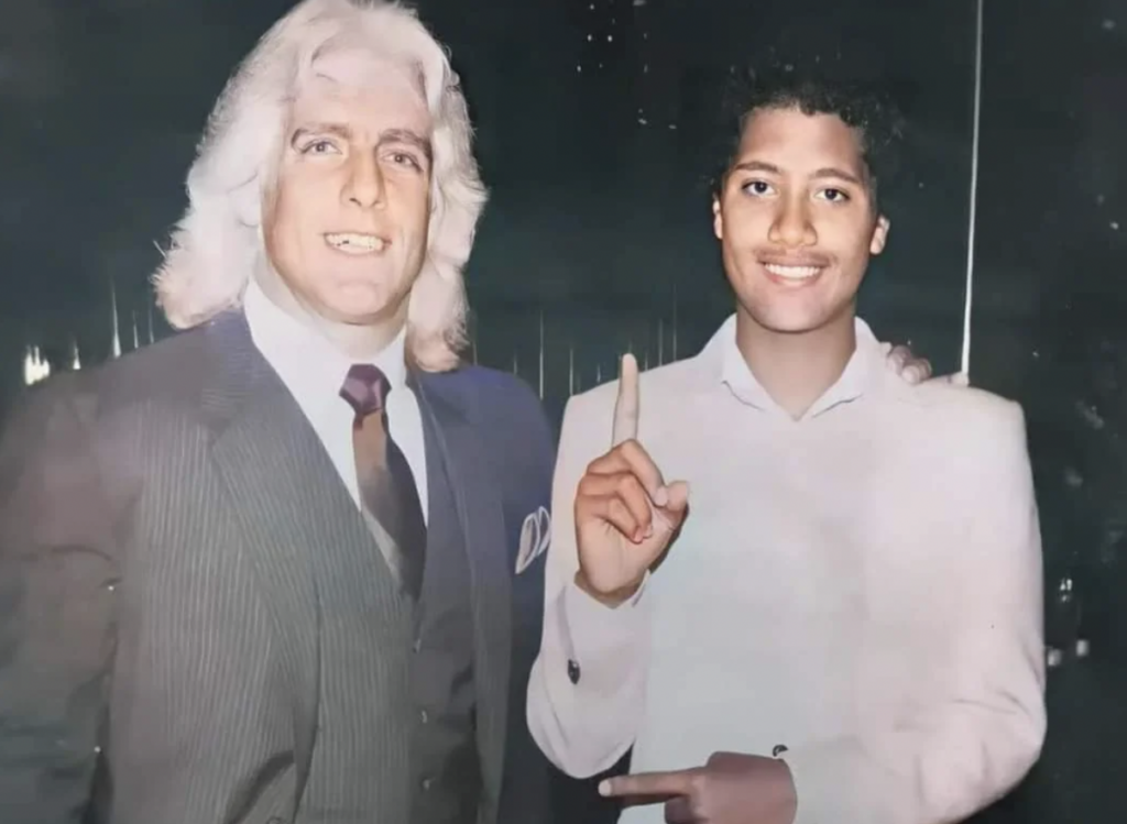 An older man with white hair wearing a gray suit, white shirt, and dark tie stands next to a young man with curly brown hair dressed in a white outfit. The young man smiles while pointing up with one finger. Both are standing indoors against a dimly lit background.