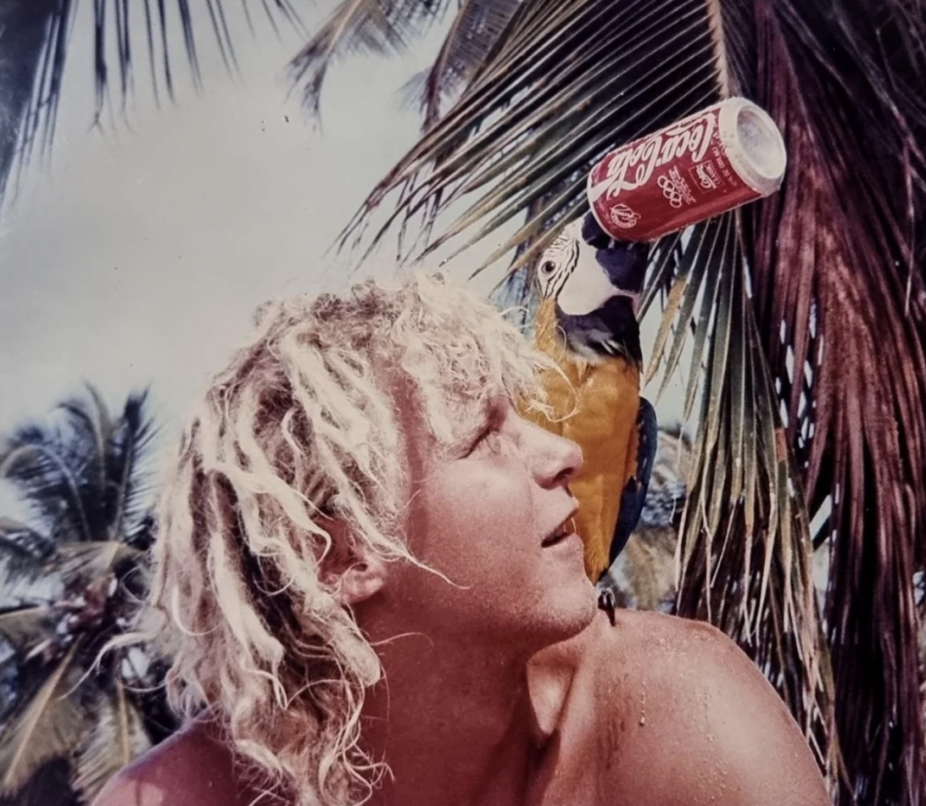 A person with curly blonde hair sits under palm trees with a colorful parrot perched on their shoulder. The parrot is holding a red Coca-Cola can in its beak. The background shows a tropical setting with clear skies and green palm fronds.
