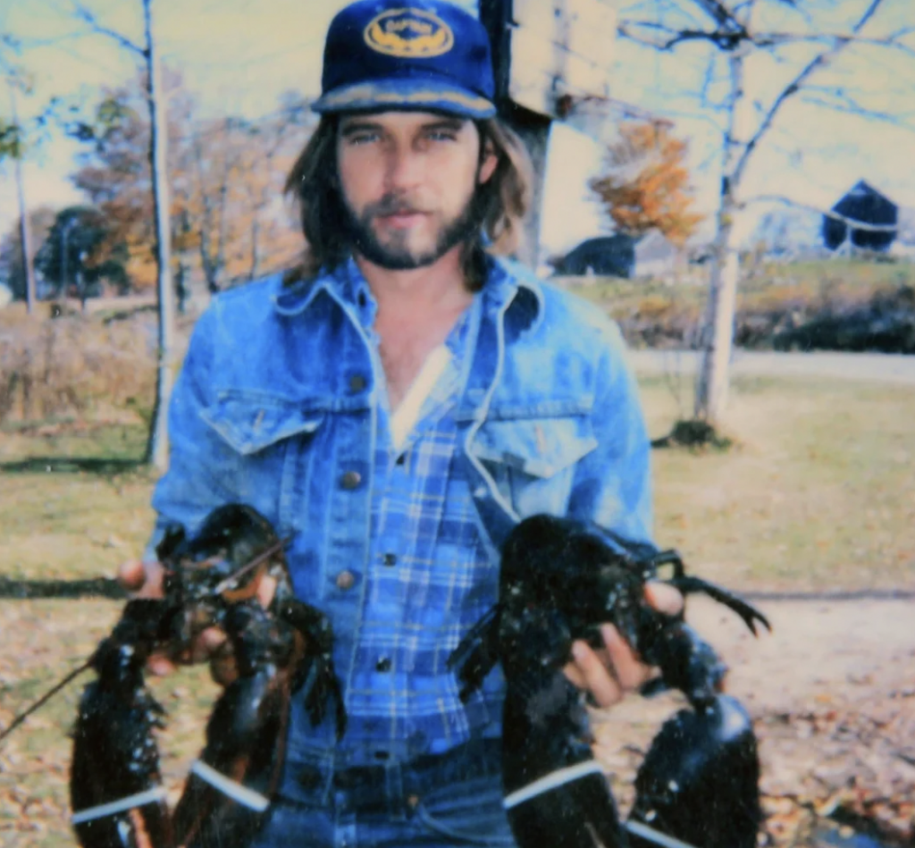 A man with long hair and a beard, wearing a blue denim jacket, a blue checkered shirt, and a blue hat with a logo, holds two large lobsters. He is standing outdoors on a sunny day with trees and a rural landscape in the background.