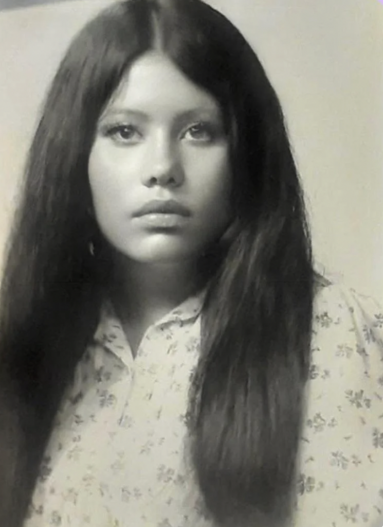 Black and white portrait of a young woman with long, straight hair, wearing a light-colored, floral patterned blouse. She gazes directly at the camera with a neutral expression.