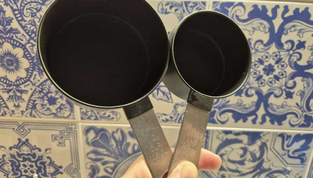 A hand holds two measuring cups, one marked 1/2 cup (125 ml) and the other marked 1/4 cup (62.5 ml), against a backdrop of blue and white patterned tiles.