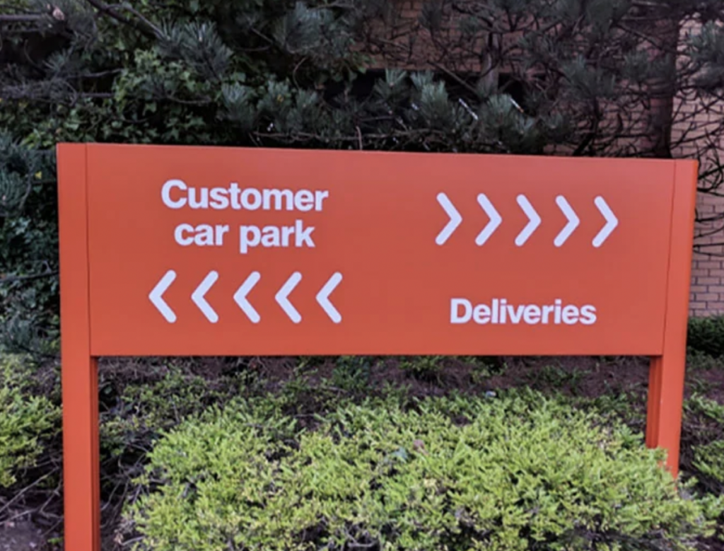 An orange sign with white text and arrows. The left side reads "Customer car park" with left-pointing arrows, and the right side reads "Deliveries" with right-pointing arrows. The sign is situated in front of greenery and a brick wall.