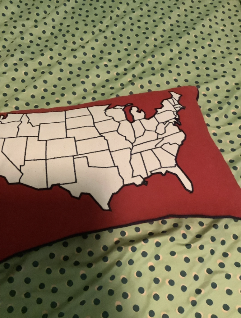 A pillow with a map of the United States drawn in white with black borders, placed on a green polka-dotted bedspread.