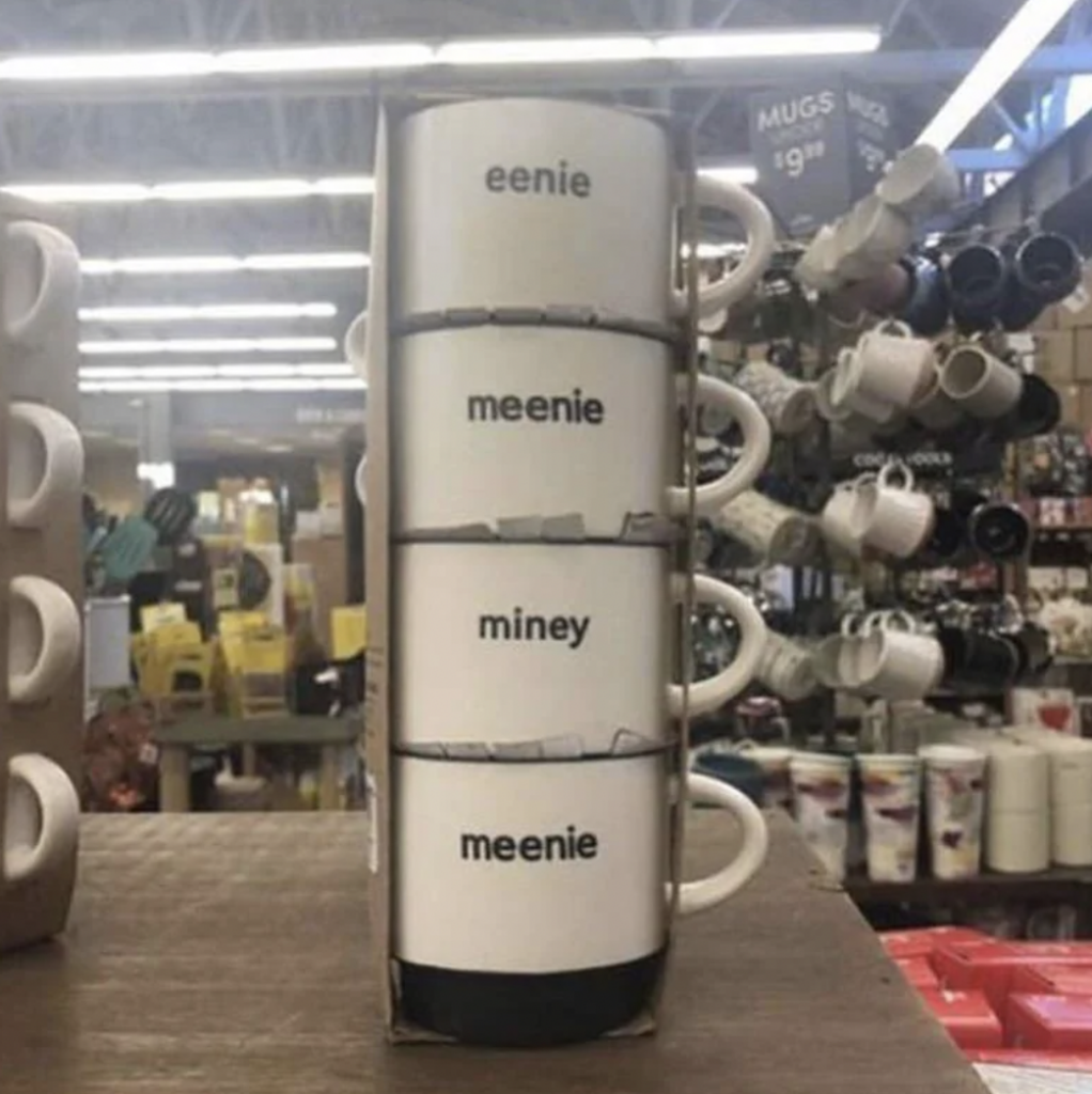 A stack of four white mugs displayed in a store, each labeled with one of the words "eenie," "meenie," "miney," and "meenie." The mugs are placed on a wooden surface, and surrounding store shelves are visible in the background.