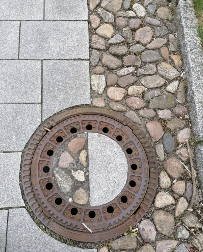 A manhole cover partially overlapping a sidewalk and a cobblestone pathway. The cover is circular and metallic with holes around the perimeter, revealing more cobblestone underneath. To the right, there is a grassy edge bordering the cobblestones.