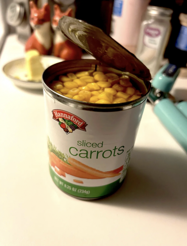 A can labeled "Hannaford Sliced Carrots" is open to reveal corn inside. The can is placed on a kitchen countertop with a teal can opener nearby, a dish with butter, and decorative fox figures in the background.
