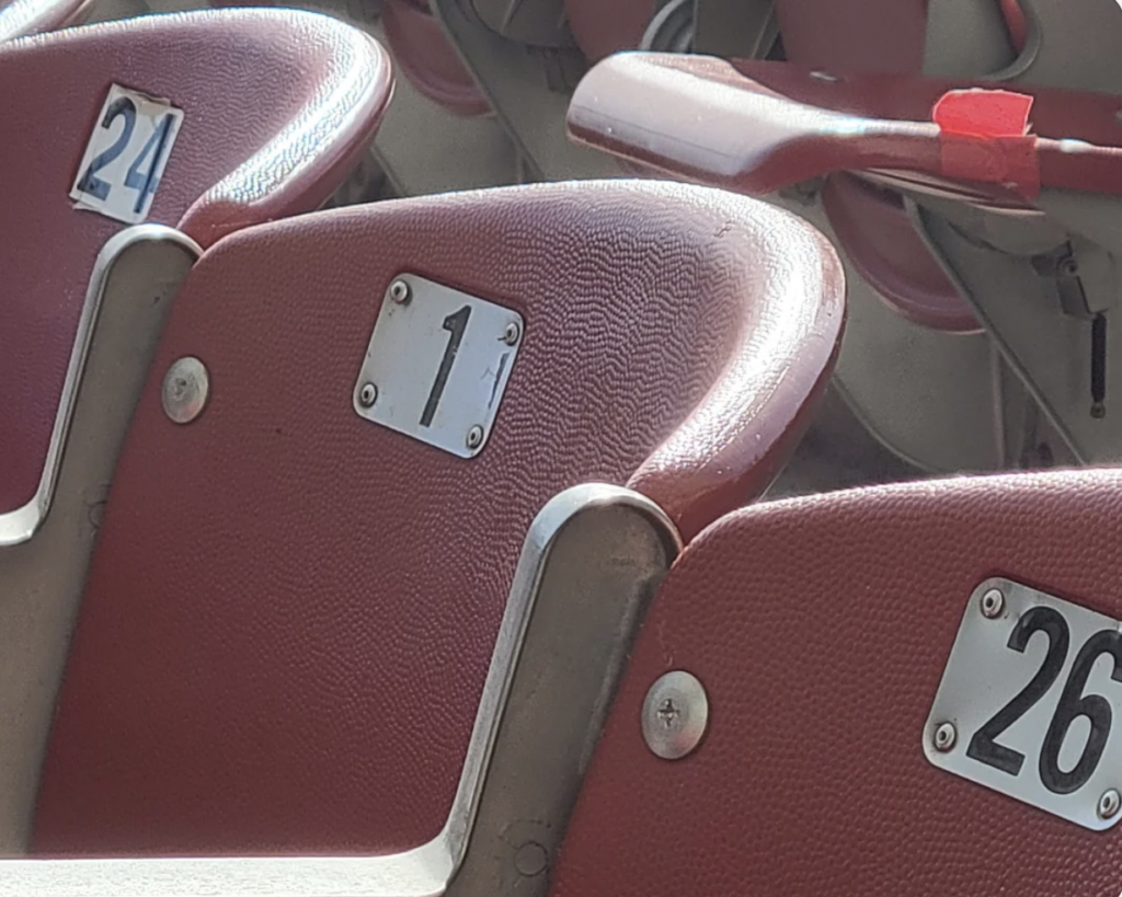 A close-up of stadium seats with numbers 24, 1, and 26 visible. The seats are red with white number plates attached to the top part of the backrests. The armrests have red tape on them. Sunlight is casting shadows over part of the seats.