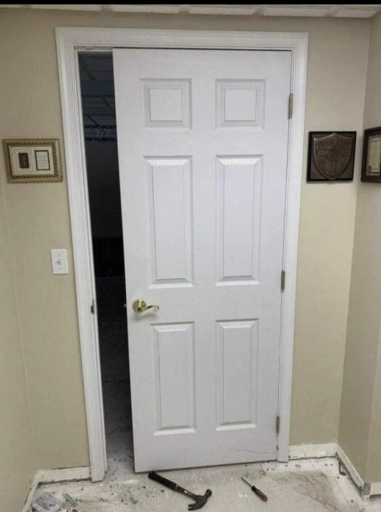 A white door installed upside down, with the doorknob unusually placed near the top. The doorframe is inside a beige-colored room with plaques on the walls. A hammer and some debris are scattered on the floor nearby.