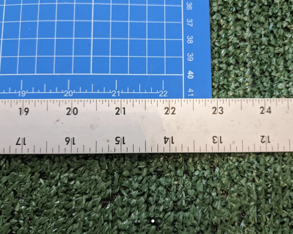 A close-up view of a ruler and a cutting mat. The ruler, marked in both inches and centimeters, is placed horizontally and measures just over 23 inches. The blue cutting mat has a grid and is placed on top of a textured green surface.