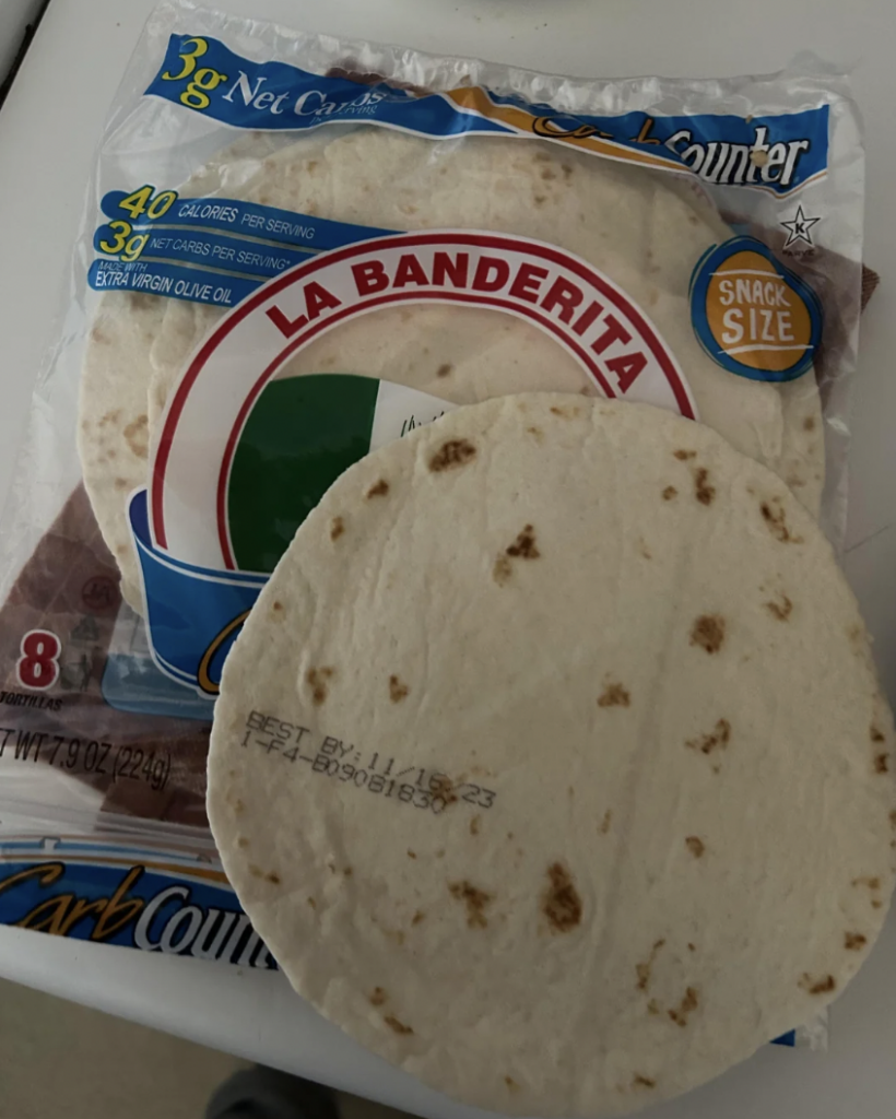 A package of La Banderita brand tortillas is shown, featuring 8 snack-size tortillas labeled as “Carb Counter” with 40 calories per serving. One tortilla is outside the package displaying the "BEST BY 11/20/23" date. The packaging is predominantly white with blue and orange accents.