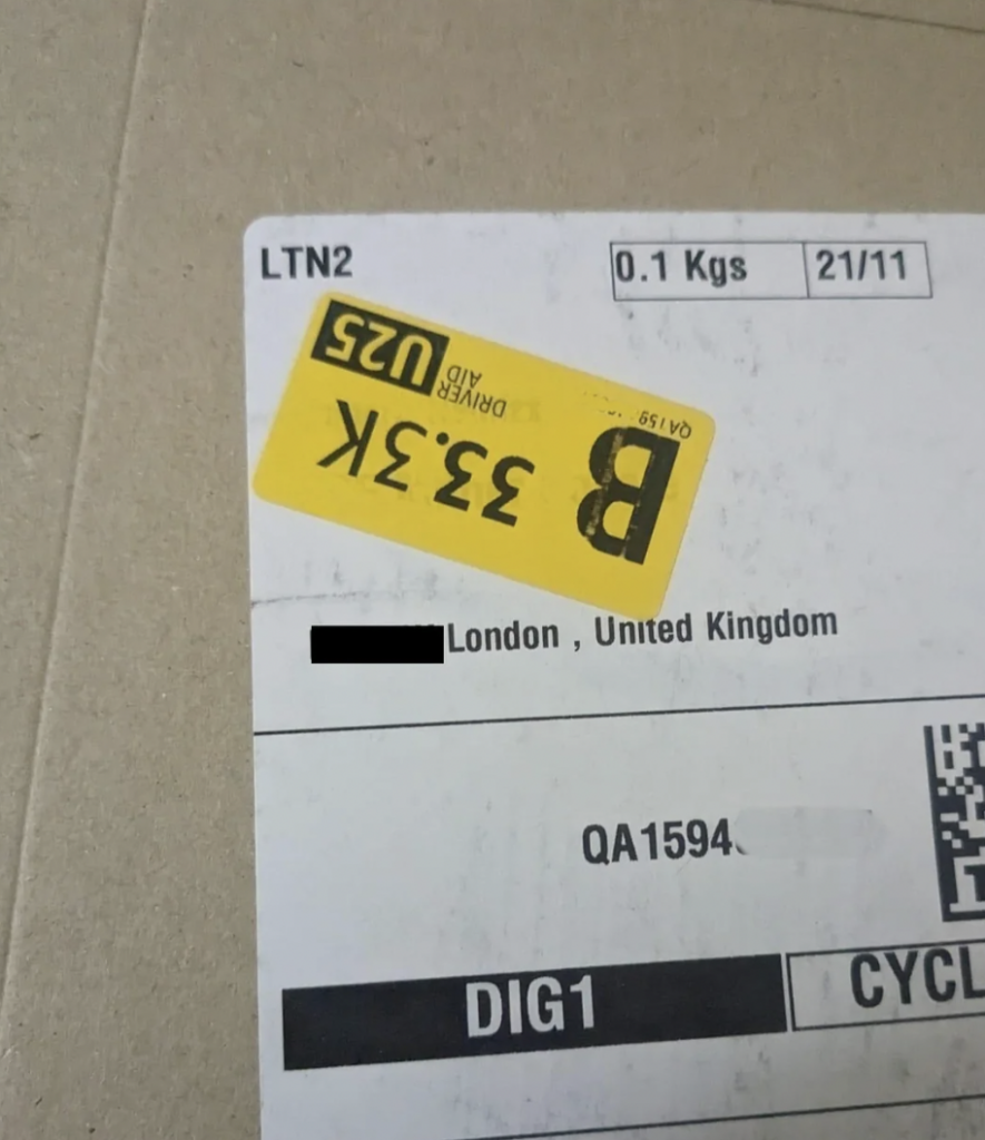 A close-up of a shipping label on a cardboard box. The label includes a yellow sticker with "UPS" and "B 333K." It also indicates the package weight as "0.1 Kgs" and displays the destination as London, United Kingdom. The tracking number is partially obscured.