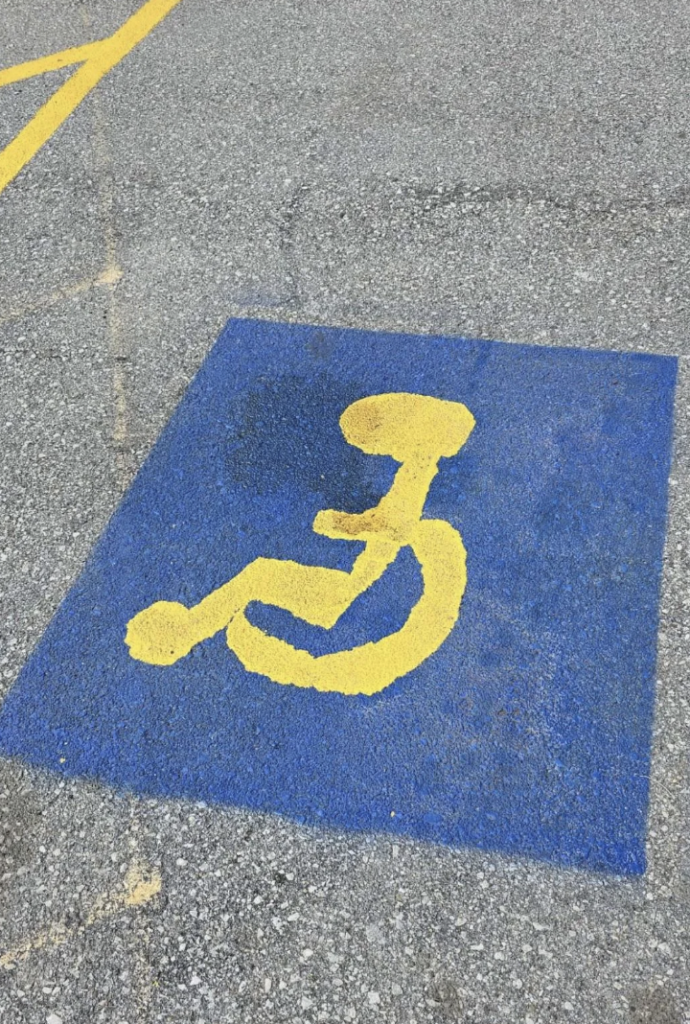 A parking space with a blue background and a yellow disabled parking symbol painted on the asphalt. The symbol signifies that the space is reserved for individuals with disabilities. The surrounding area shows a rough, textured grey surface.