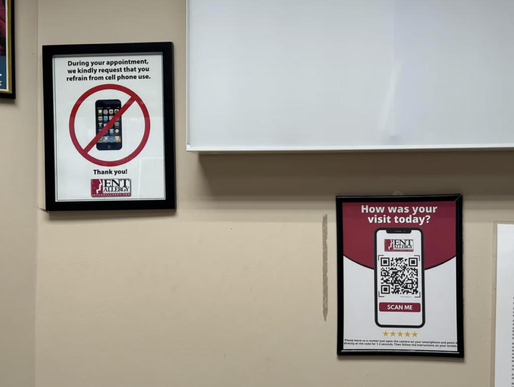 Two signs hang on a wall beneath a whiteboard. The left sign requests no cell phone use during appointments, with an image of a crossed-out phone. The right sign inquires about the visit experience with a QR code labeled "Scan Me" for feedback submission.