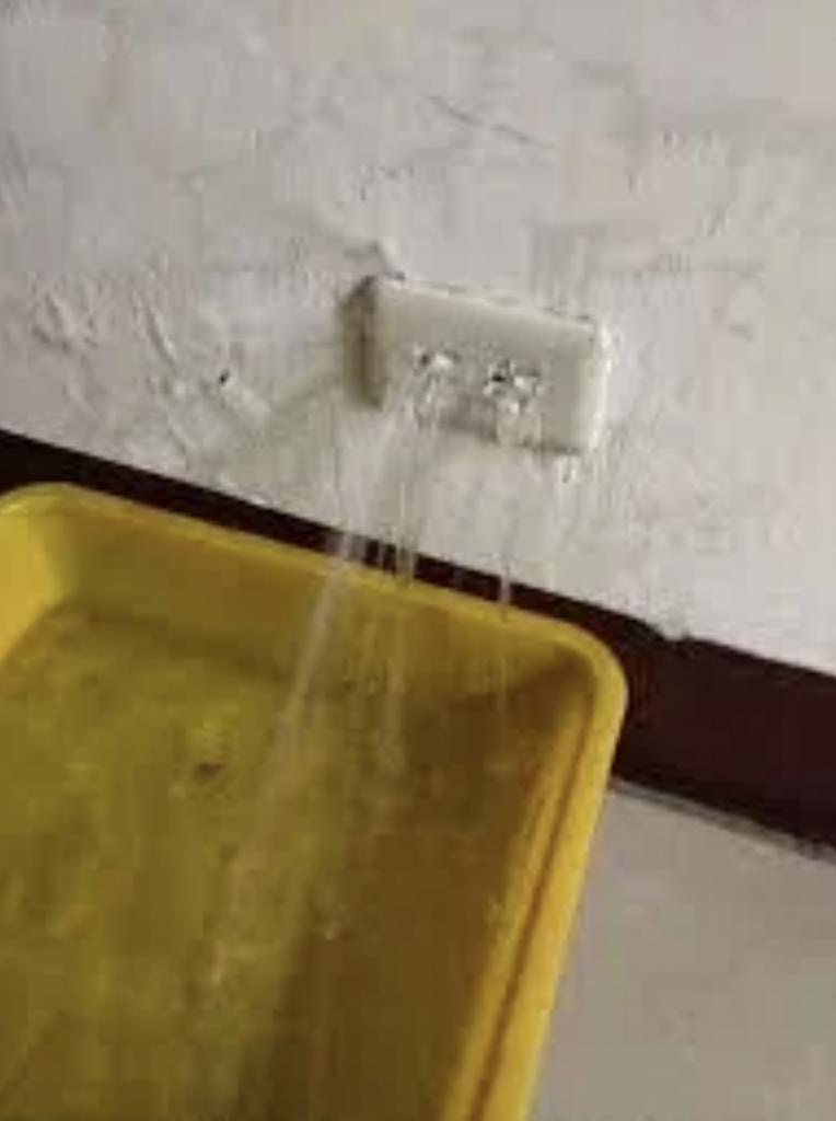 A yellow container is placed below a leaking electrical outlet on a wall. Water is streaming out of the outlet and into the container.