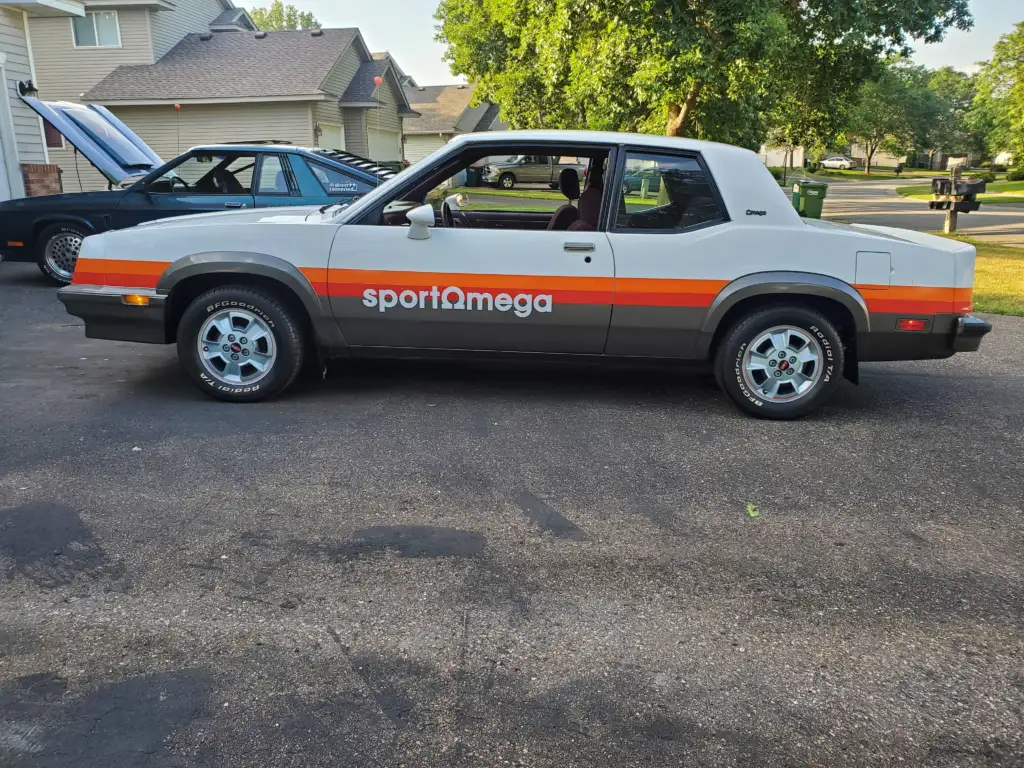 A white two-door coupe with "sportomega" decals in orange and gray parked in a residential driveway. Another black car is seen in the background. The scene includes houses, trees, and a sunny day.