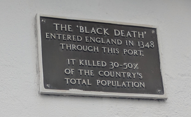 A metal plaque on a white wall reads: "The 'Black Death' entered England in 1348 through this port. It killed 30-50% of the country's total population.