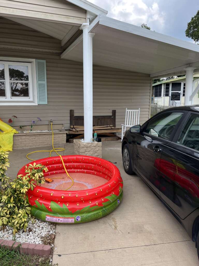 A red inflatable kiddie pool is set up on a concrete driveway next to a black car in front of a house with a covered porch. The porch has a white rocking chair and some potted plants. A yellow hose is connected to the pool, and some shrubs are in the foreground.