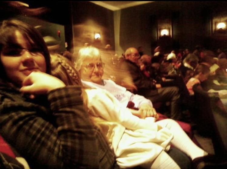 An audience sits in a dimly lit theater. In the foreground, a woman with dark hair and a checkered jacket rests her chin on her hand, while an elderly woman with glasses, a white sweater, and light purple blouse sits next to her. The background shows other audience members.