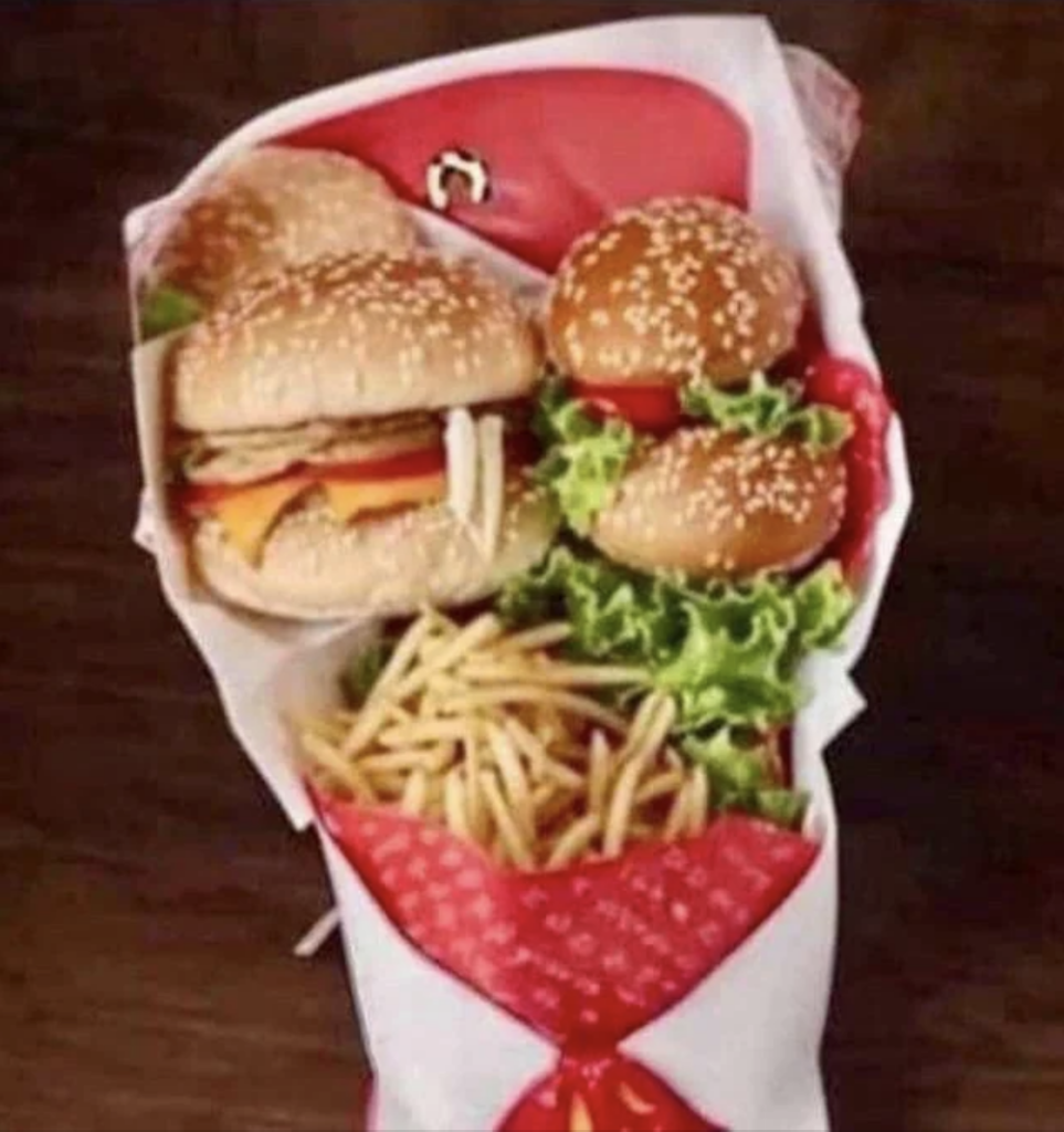 A bouquet-style arrangement featuring two cheeseburgers with lettuce, tomato, and sesame seed buns, accompanied by a serving of french fries, all wrapped in red and white paper with a ribbon design. The background is a dark wooden table.