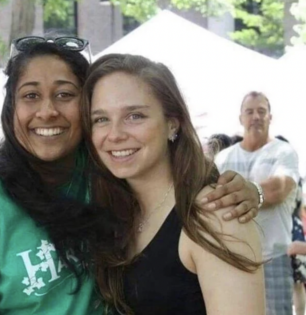 Two women, one in a green shirt and the other in a black tank top, smile as they pose for a photo outdoors. The woman in the green shirt has her hand around the other woman's shoulder. A man in a white plaid shirt stands in the background, smiling.