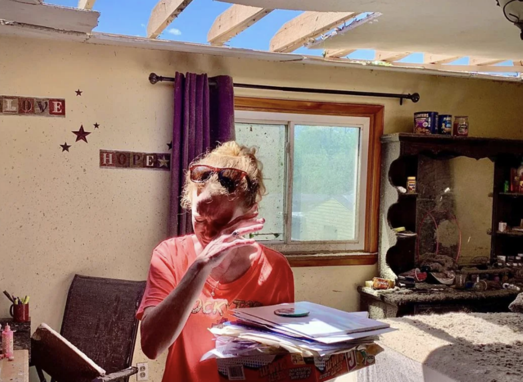 A person stands inside a damaged house with a partially collapsed ceiling and scattered debris. They are holding a pile of papers and mail, wearing sunglasses, and covering their face with one hand. Sunlight filters through the damaged roof, illuminating the scene.