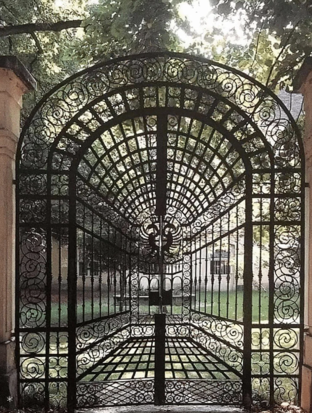 A tall, intricately designed iron gate is adorned with ornate patterns and a central emblem. The gate, set between two stone pillars, opens to a green lawn with sunlight filtering through the surrounding trees.
