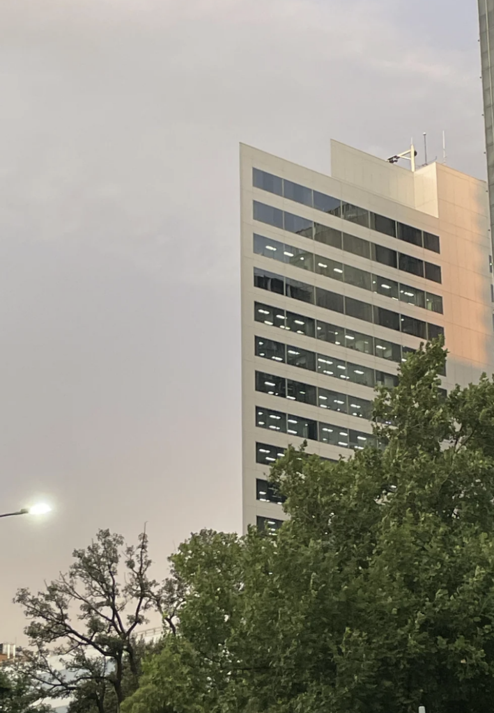 The image shows a modern high-rise building with a sleek, angular design, featuring large windows reflecting the sky. In the foreground, a densely foliated tree partially obscures the building. The sky has a soft pastel hue, indicating early morning or evening.