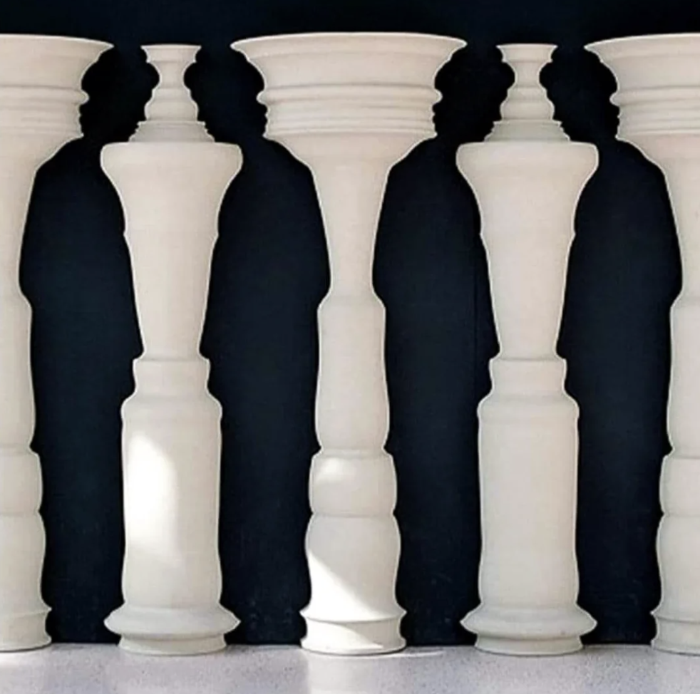 A visual illusion where five white columns are placed against a black background. The columns have unique shapes, and the negative space between them appears to form five human figures standing in a row.