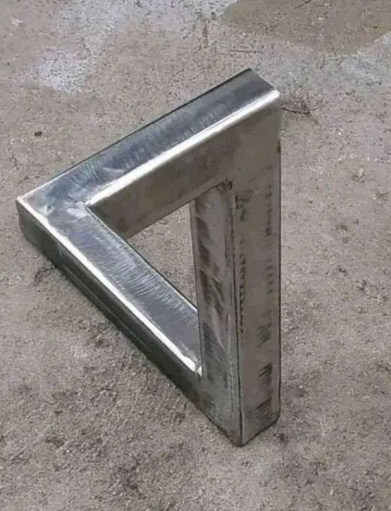 A metal sculpture appears to form an impossible triangle, an optical illusion where the structure seems to bend in a way that defies the laws of geometry. The sculpture is placed on a rough, gray, concrete surface.