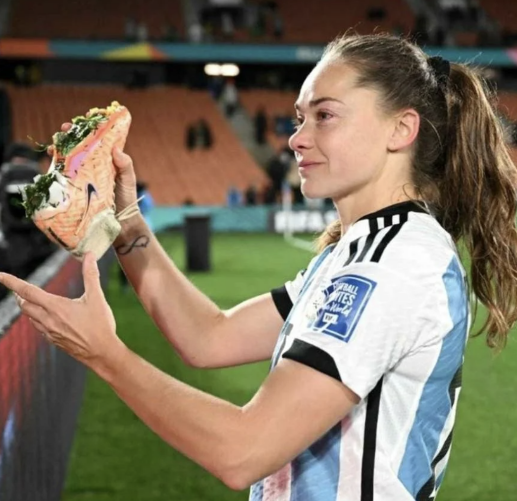 A soccer player wearing a light blue and white striped jersey holds up a shoe filled with what appears to be food. She is on a soccer field with stadium seats and spectators in the background. The player has long hair tied back and a determined expression.