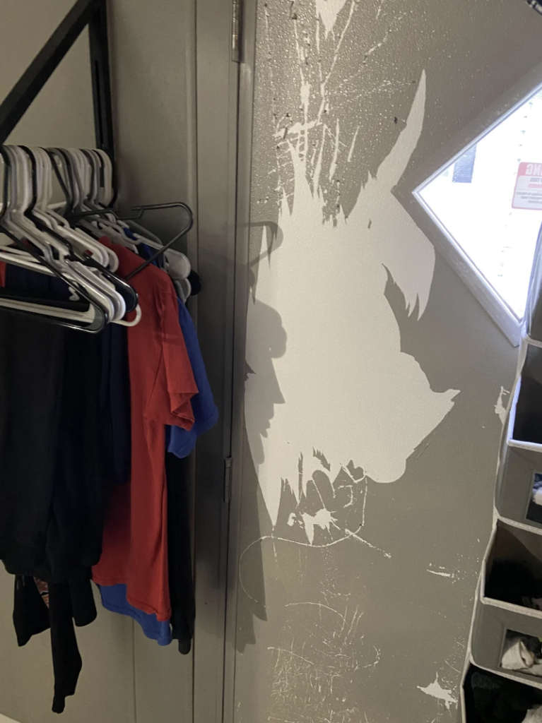 A closet showing an assortment of clothes hung on hangers to the left. The wall on the right has visible scratches and wear, creating a white, abstract pattern. The door has a small diamond-shaped window, and part of a shelving unit with small cubbies is visible on the right.