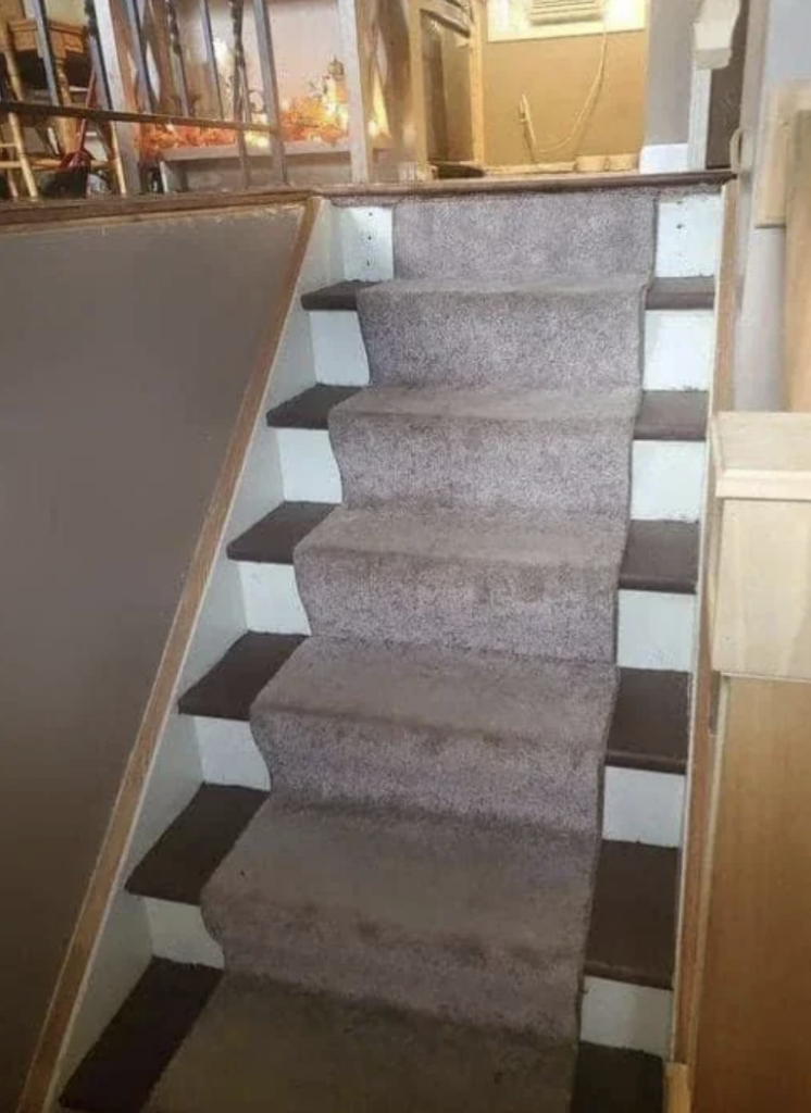 A staircase with alternating carpet-covered steps in a living space. The steps are unusually shaped, with some pairs of steps wider than others, creating an uneven pattern. Railings and part of a kitchen are visible in the background.