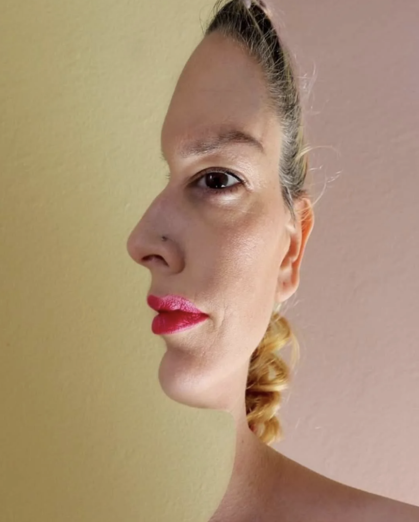 A woman stands against a two-tone wall with her face positioned in a way that creates an optical illusion, making it appear as if part of her face, including one eye, an eyebrow, and lips with bright pink lipstick, merges with the wall.