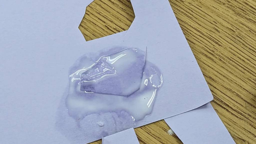 A piece of white paper with a hole sits on a wooden surface, with a translucent, slightly purple liquid spilled on it. The liquid has spread into an irregular shape, soaking into the paper.