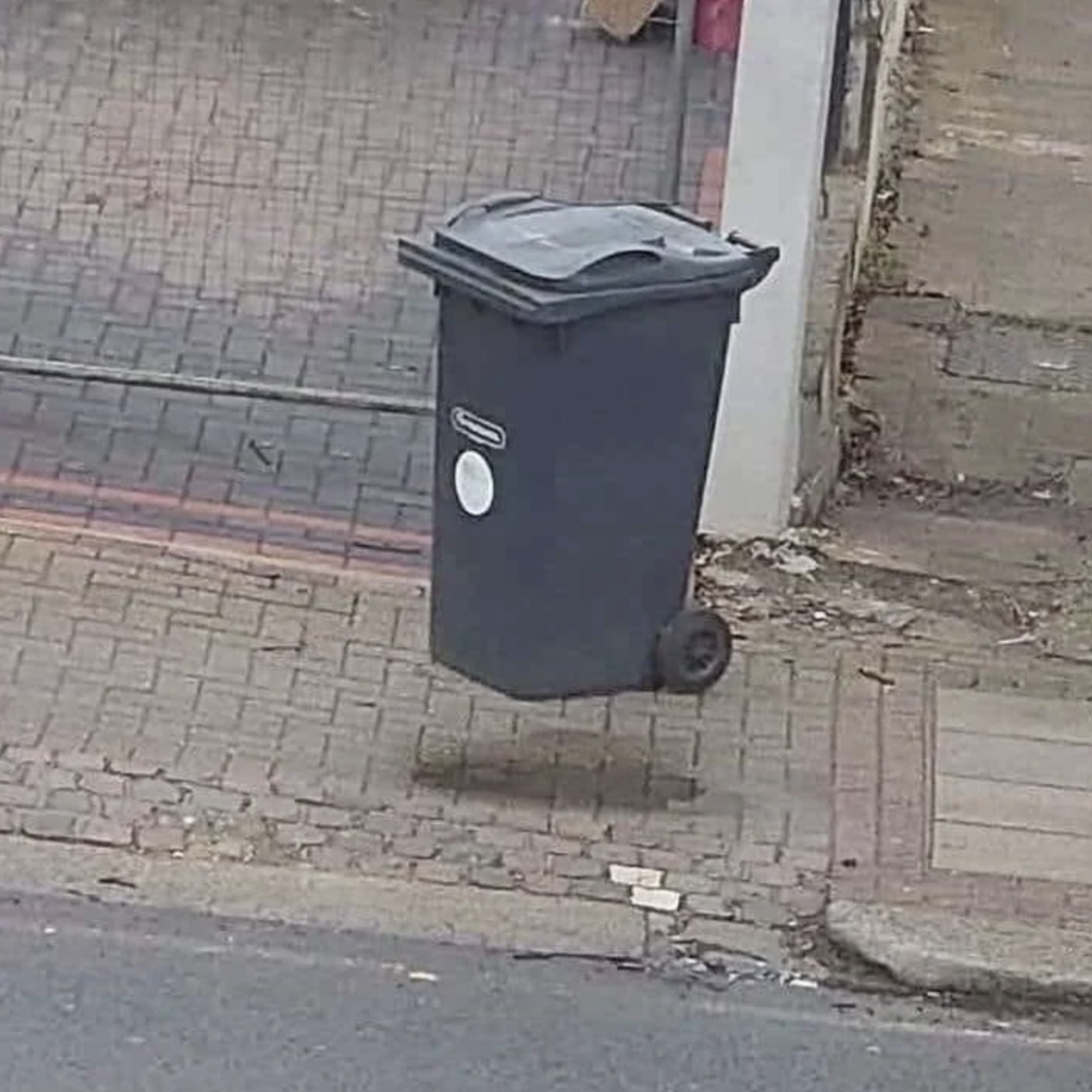 A black wheeled trash bin appears to be floating above the ground on a paved pathway next to a driveway. The bin casts a distinct shadow beneath it, creating an illusion of being airborne.